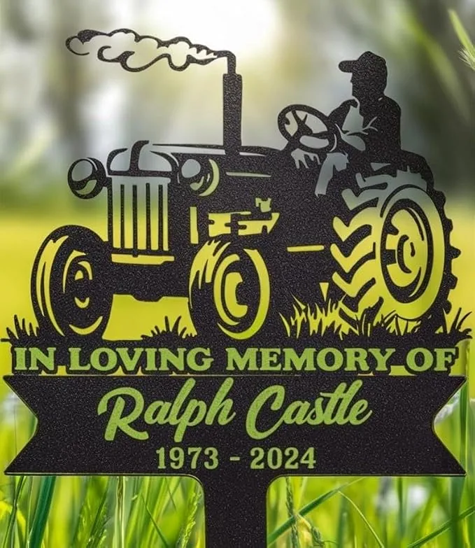 Metal memorial garden sign showing a tractor silhouette and driver with text “In Loving Memory of Ralph Castle 1973 - 2024”, staked in grassy outdoor setting