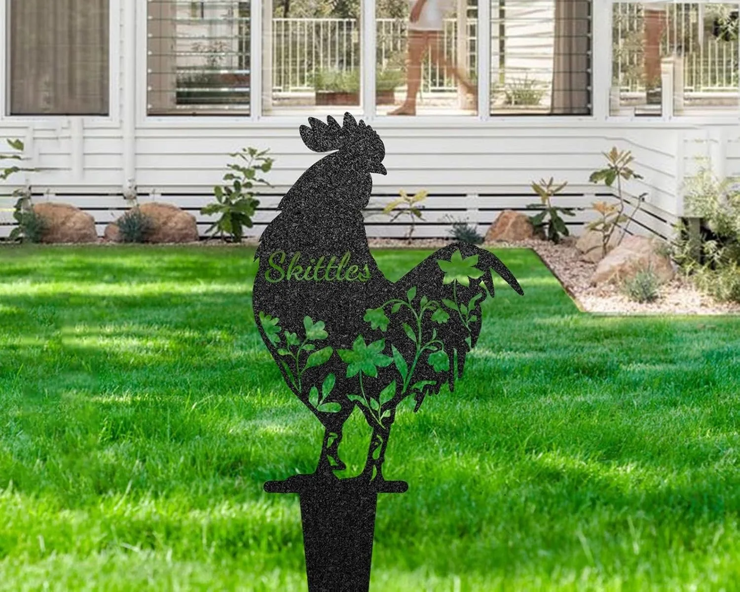 Black metal rooster yard stake with floral cutouts and the name “Skittles” displayed, planted in a green lawn in front of a white house