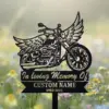 Black metal memorial stake in a flower field depicting a winged motorcycle and text reading "In loving Memory Of" with placeholder "CUSTOM NAME" and years "1955-2025"