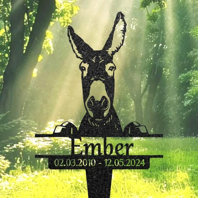 Black metal donkey memorial sign reading "Ember" with dates 02.03.2010 - 12.05.2024 set against a sunlit forest background