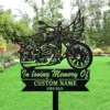 Black metal memorial garden sign shaped like a winged motorcycle reading 'In loving Memory Of', 'CUSTOM NAME', and '1955-2025', staked in a green lawn with trees and flowers in background.