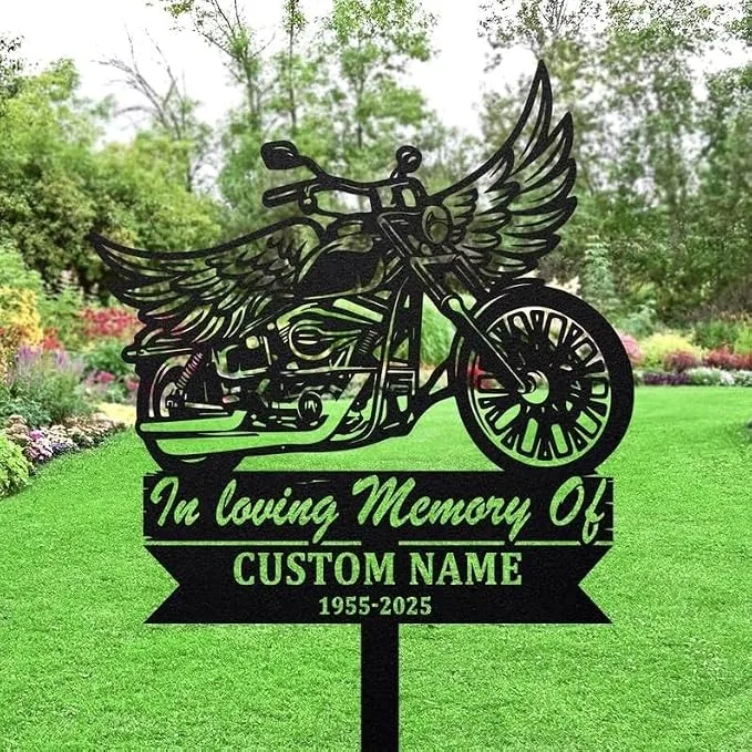 Black metal memorial garden sign shaped like a winged motorcycle reading 'In loving Memory Of', 'CUSTOM NAME', and '1955-2025', staked in a green lawn with trees and flowers in background.