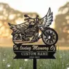 Black metal memorial yard sign shaped like a winged motorcycle reading "In loving Memory Of" with placeholder "CUSTOM NAME" and dates, placed in grass at sunrise.