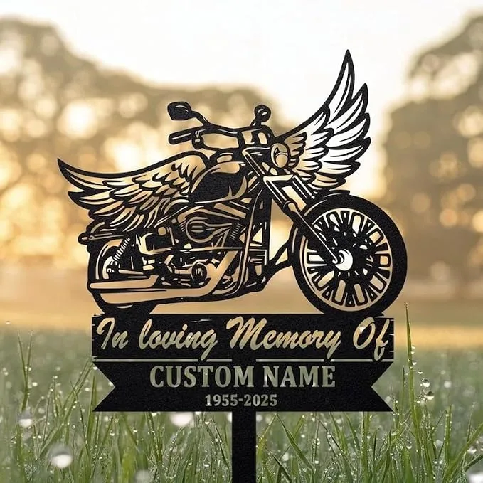 Black metal memorial yard sign shaped like a winged motorcycle reading "In loving Memory Of" with placeholder "CUSTOM NAME" and dates, placed in grass at sunrise.