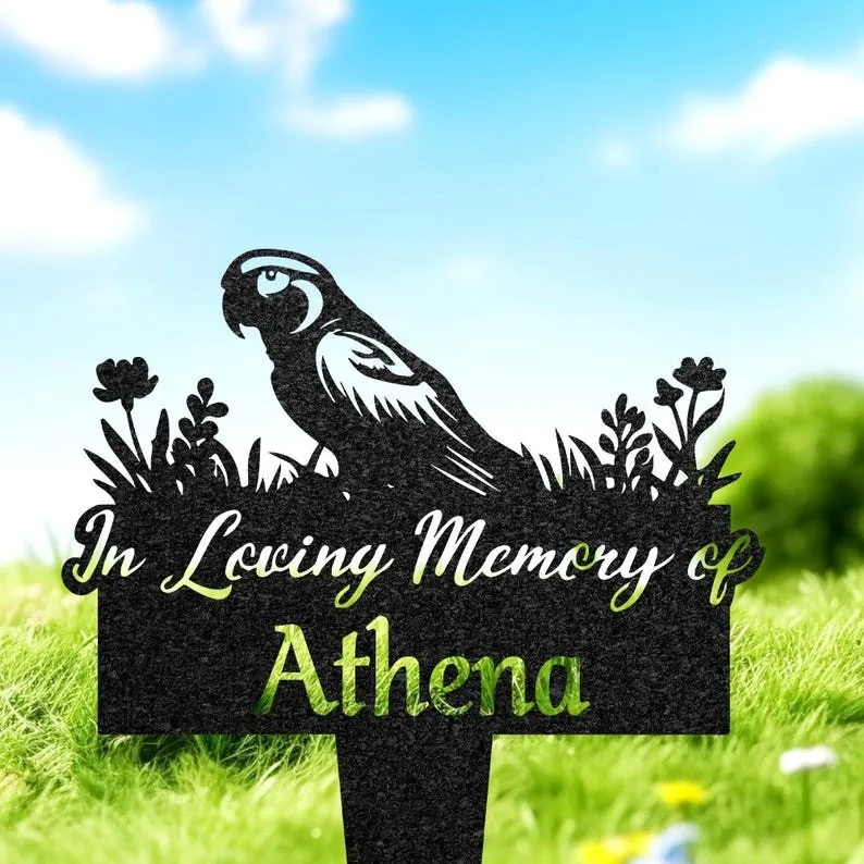 Black garden memorial stake with parrot silhouette and flowers reading "In Loving Memory of Athena" placed in green grass under blue sky