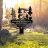 Black metal memorial sign shaped like a cross with trees, a hunter aiming at deer, angel wings and the text “Hunting in Heaven Russel Smith 1963 - 2025,” placed outdoors on grass near a wooded pond at sunrise.