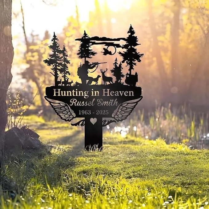 Black metal memorial sign shaped like a cross with trees, a hunter aiming at deer, angel wings and the text “Hunting in Heaven Russel Smith 1963 - 2025,” placed outdoors on grass near a wooded pond at sunrise.
