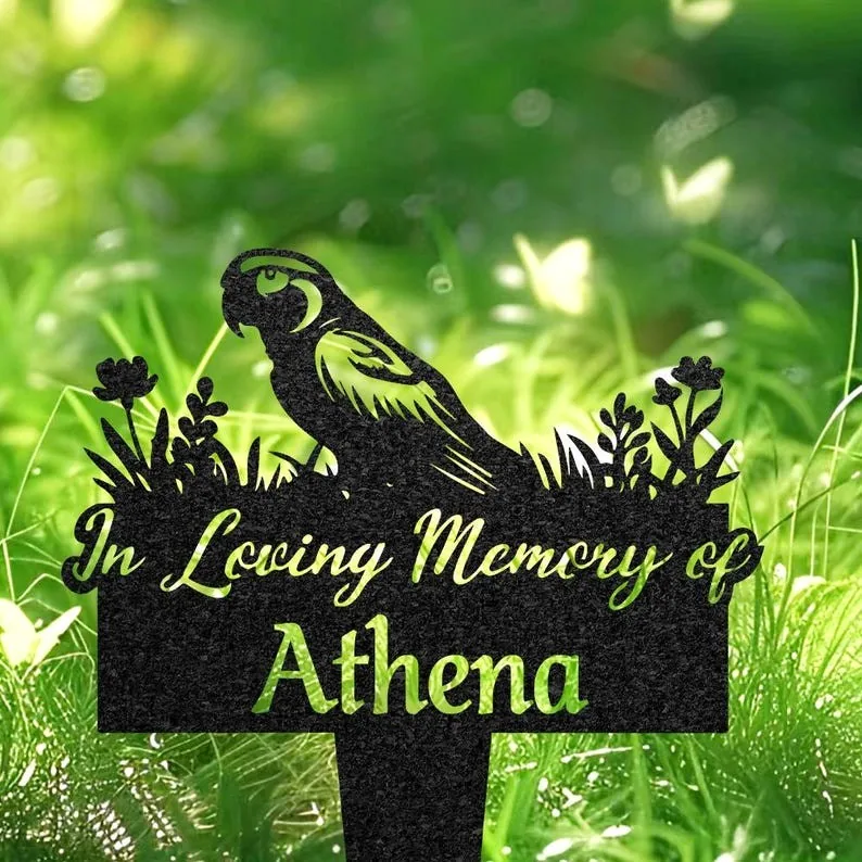 Black memorial garden stake with a parrot silhouette and floral accents reading "In Loving Memory of Athena" placed in grassy outdoor setting