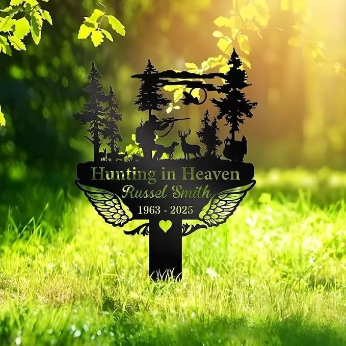 Black metal memorial sign reading "Hunting in Heaven" with name "Russel Smith" and dates "1963 - 2025," silhouette of a hunter, deer, trees and angel wings staked in sunlit grass