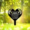 Black heart-shaped memorial stake in sunlit green park reading "In loving memory SON" and personalized name and dates