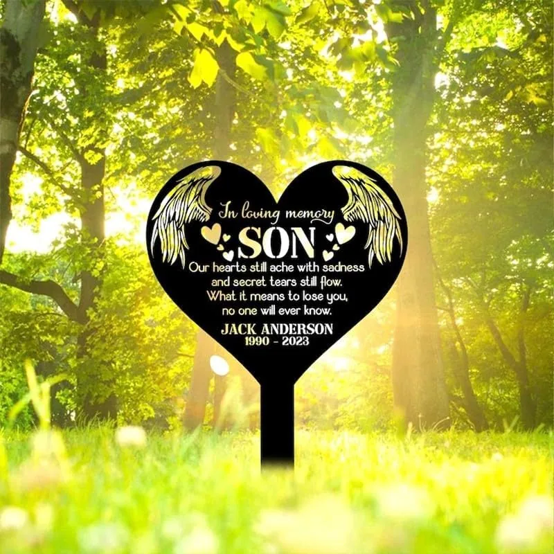 Black heart-shaped memorial stake in sunlit green park reading "In loving memory SON" and personalized name and dates