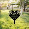 Black heart-shaped memorial yard stake reading "In loving memory SON" with angel wings and personalized name/dates, placed in a sunny grassy park near picnic tables