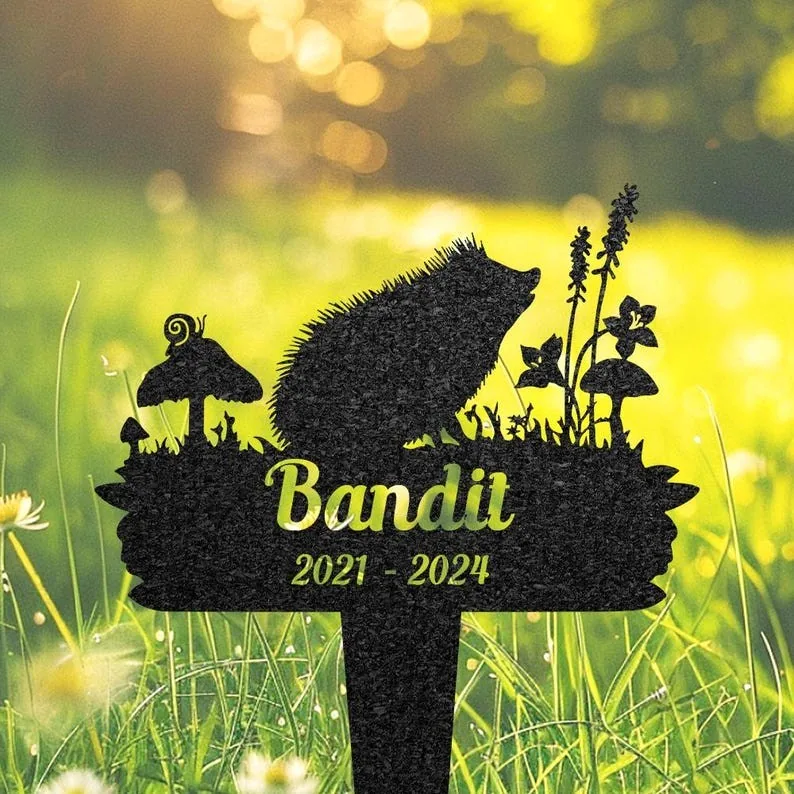 Black garden memorial stake silhouette of a hedgehog among mushrooms and flowers with name 'Bandit' and dates '2021 - 2024' placed in sunlit grassy lawn