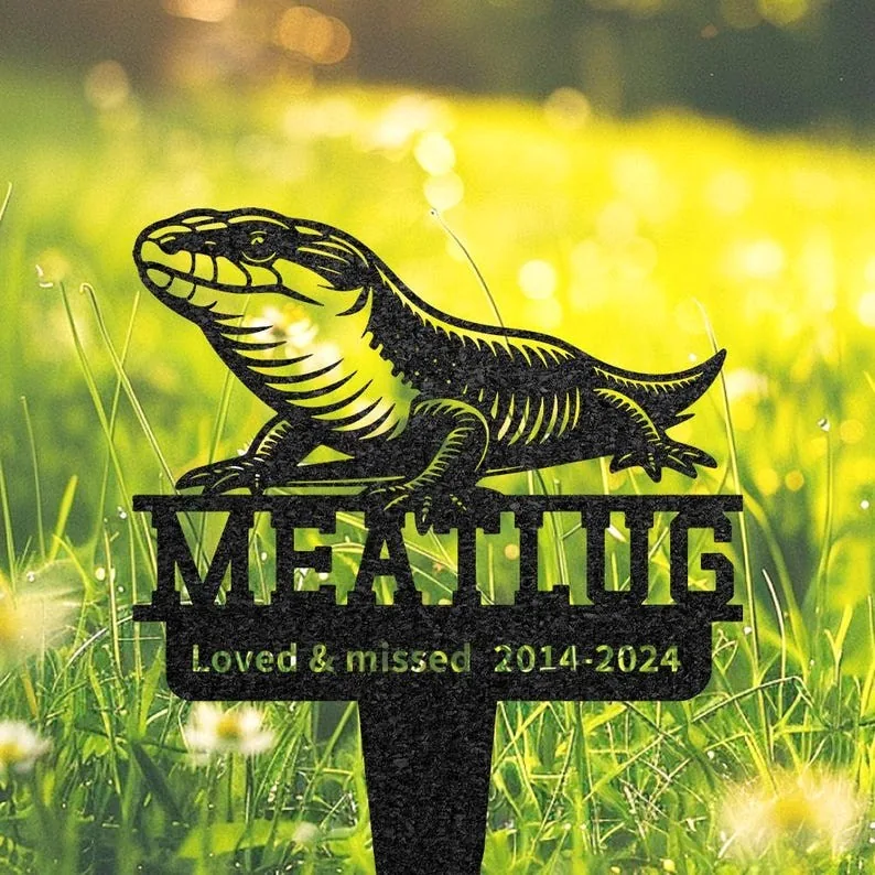 Black metal garden memorial stake shaped like a lizard with the name "MEATLOAF" and text "Loved & missed 2014-2024" set in sunlit grassy yard