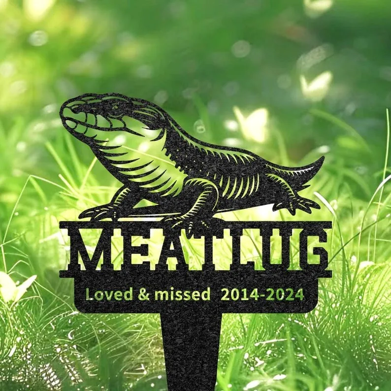 Black metal garden memorial stake showing a lizard above the name "MEATLOAF" with inscription "Loved & missed 2014-2024" stuck in green grass