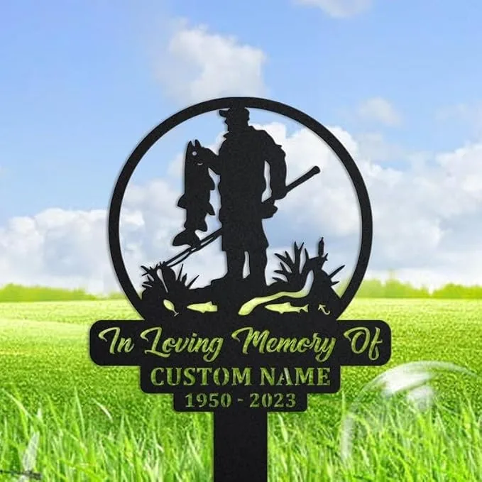 Black metal memorial garden stake showing a fisherman holding a fish inside a circle, with text reading 'In Loving Memory Of', 'CUSTOM NAME' and years '1950 - 2023', placed in grassy outdoor setting