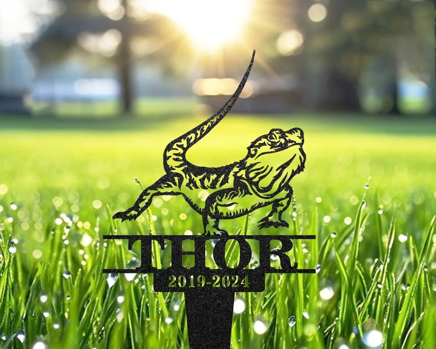Black metal garden stake shaped like a lizard with the name THOR and dates 2019-2024 set in sunlit dewy grass