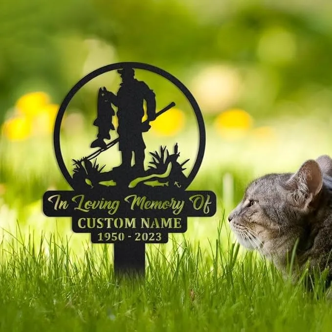Black metal garden memorial stake showing a silhouette of a fisherman holding a fish, text reading "In Loving Memory Of" and placeholder "CUSTOM NAME 1950 - 2023" staked in grass with a cat nearby