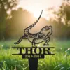 Black metal garden memorial stake shaped like a bearded dragon above the name THOR with dates 2019-2024 in grassy yard at sunrise