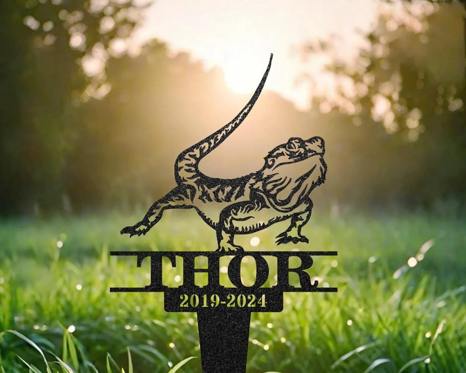 Black metal garden memorial stake shaped like a bearded dragon above the name THOR with dates 2019-2024 in grassy yard at sunrise