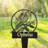 Black metal garden sign with a perched bird inside a circle and the name "Ophelia" in front of a sunlit grassy park