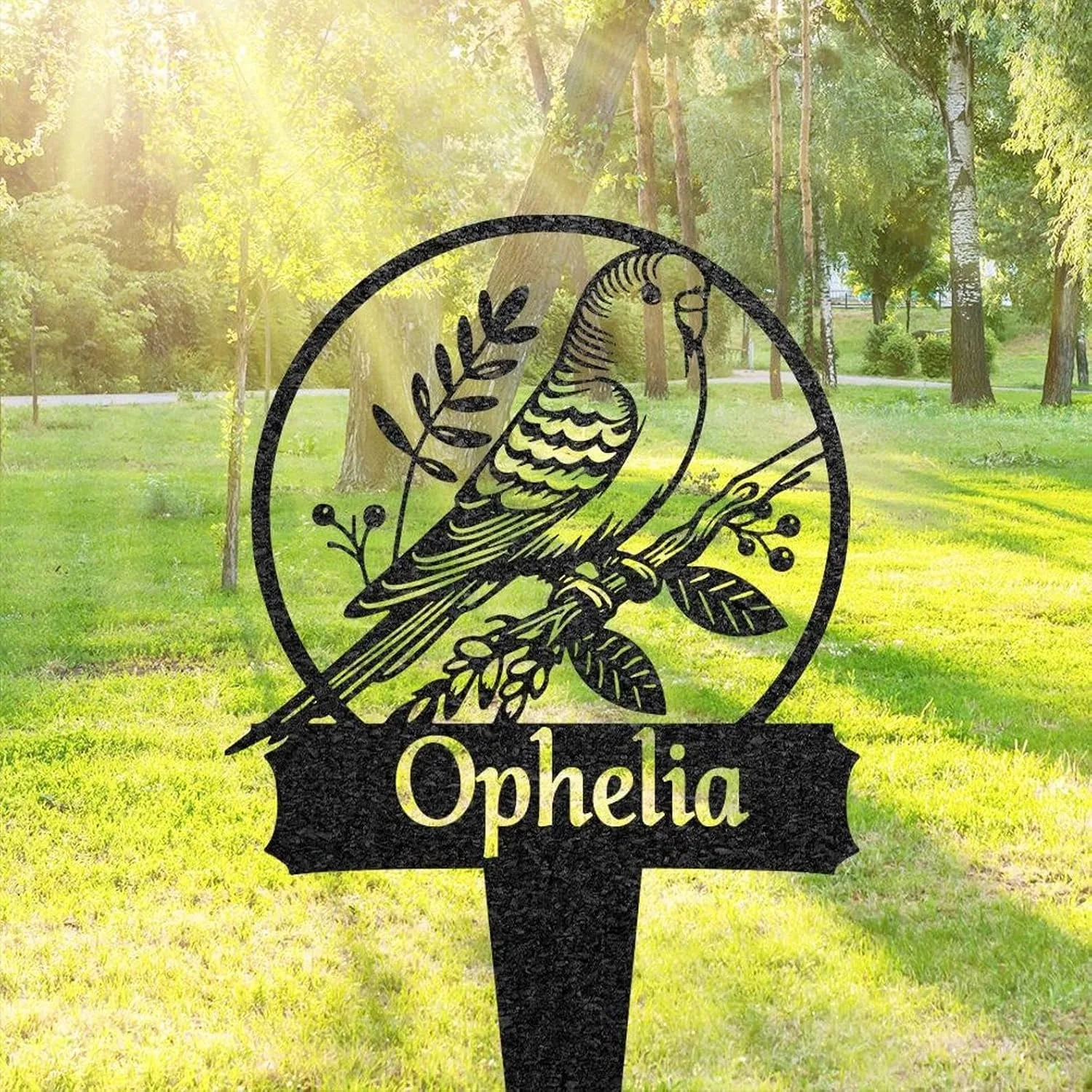 Black metal garden sign with a perched bird inside a circle and the name "Ophelia" in front of a sunlit grassy park