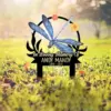 Memorial yard sign with a blue dragonfly illustration and flowers reading "In Loving Memory Of Andy Mandy 1986 - 2024" staked in grassy sunlit lawn