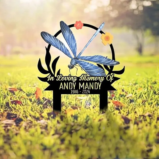 Memorial yard sign with a blue dragonfly illustration and flowers reading "In Loving Memory Of Andy Mandy 1986 - 2024" staked in grassy sunlit lawn