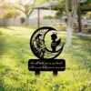 Black metal memorial lawn stake showing a praying figure holding a child beside a crescent moon and angel silhouette with text plaque reading “We will hold you in our hearts until we can hold you in our arms”