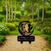 Black garden memorial stake showing Mary holding a child and angel silhouette with name and dates, placed on a gravel path amid flowering garden and birch trees