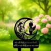 Black memorial garden stake showing Jesus holding a child inside a crescent moon and a silhouetted angel figure labeled "Baby Andy 2024-2025," placed outdoors among green grass and pink hydrangea blooms