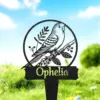 Black metal garden stake with a perched bird inside a circular frame and the name "Ophelia" displayed below, shown in grassy outdoor setting