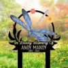 Memorial garden stake with a blue dragonfly illustration, floral accents and text reading "In Loving Memory Of ANDY MANDY 1986 - 2024" placed in a grassy flower-filled yard