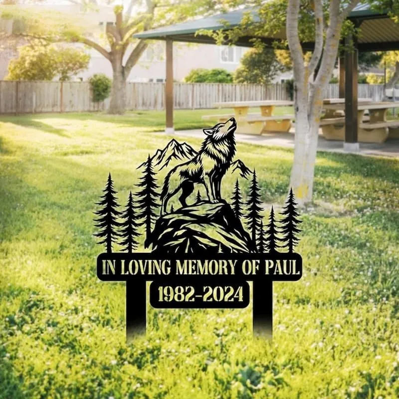 231-2 Black metal memorial yard sign with a howling wolf, pine trees and mountains reading "In Loving Memory of Paul 1982-2024" staked in a sunny grassy park near a picnic shelter