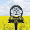 231-3 Black metal garden memorial stake shaped like a smiling hedgehog above a banner reading "In Memory Of Tito" with dates Nov 5 2018 - May 2024 against a yellow flower field and cloudy sky