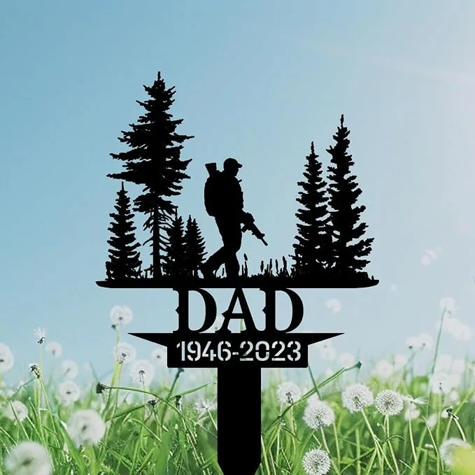 Black metal memorial garden stake silhouette of a hunter with rifle among pine trees reading “DAD 1946-2023” against a blue sky and dandelion field