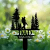 Black metal memorial garden stake reading "DAD 1946-2023" with silhouette of a soldier and pine trees among daisies in a sunny grassy background