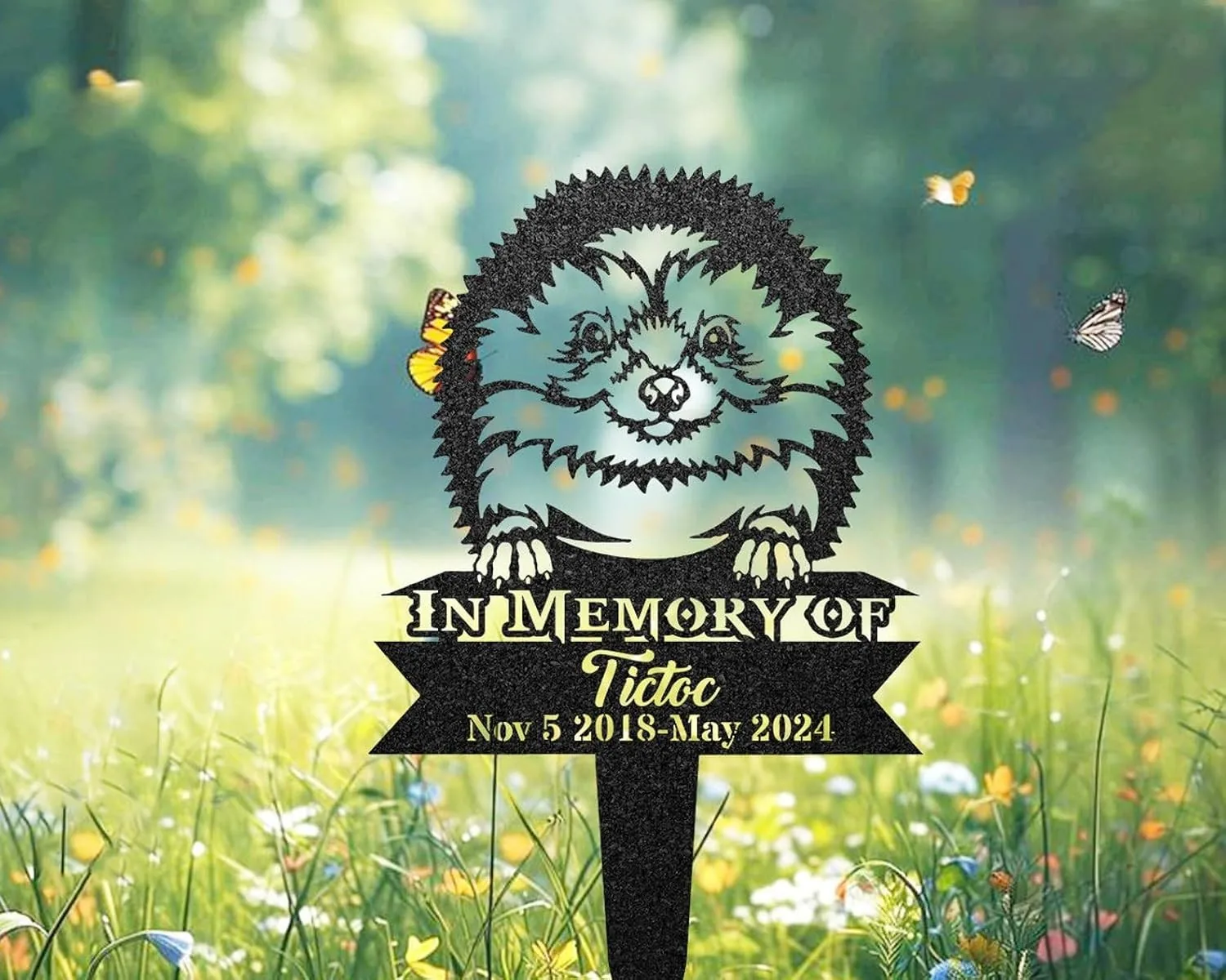 232-3 Black metal yard memorial sign with a smiling fluffy dog illustration, text “In Memory Of Tictoc” and dates “Nov 5 2018 - May 2024” staked in a sunlit meadow