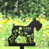 Black metal silhouette dog garden memorial stake with floral cutouts reading “Luna 2012-2024” against a blurred wildflower meadow background