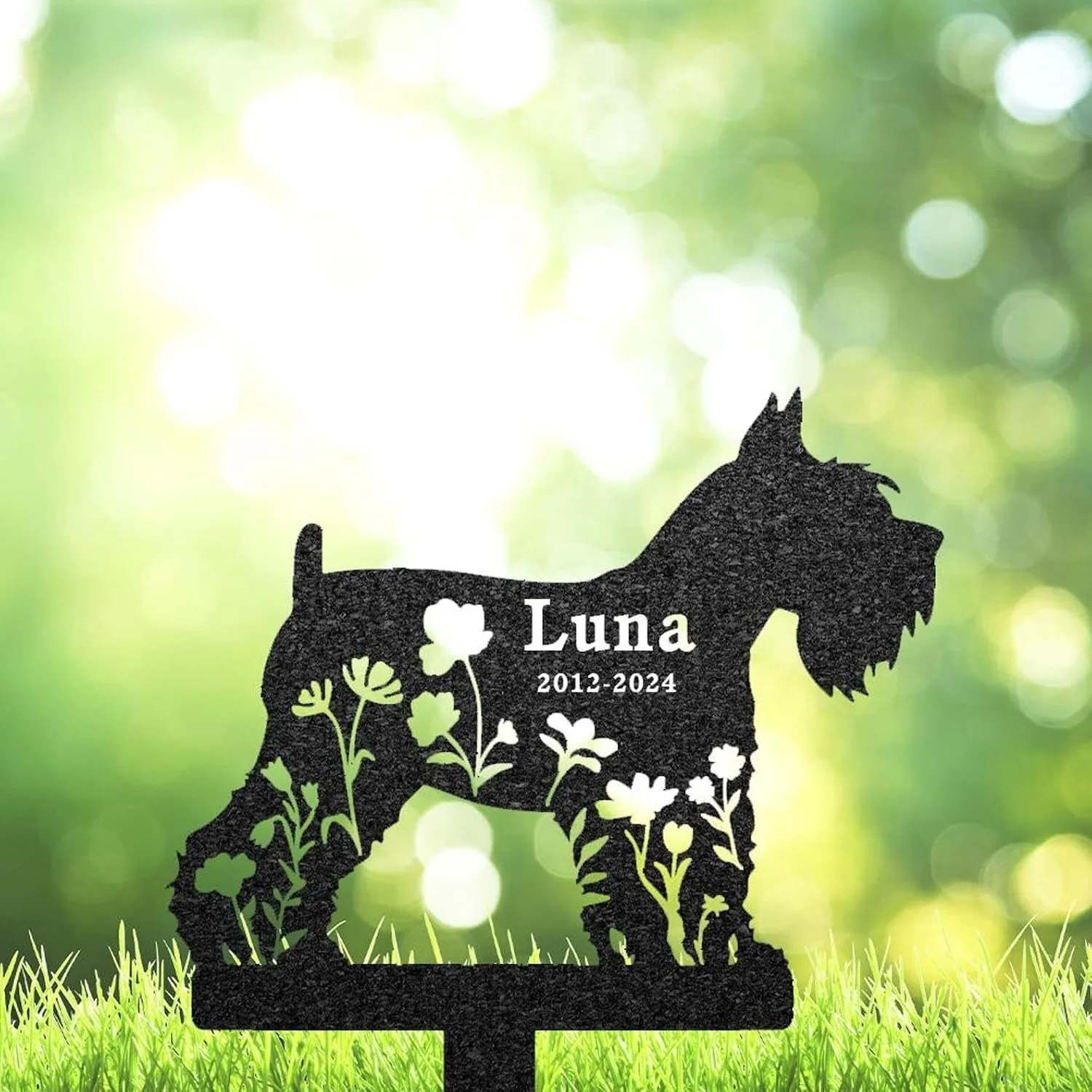 Black silhouette garden memorial stake in shape of a small dog with floral cutouts and text “Luna 2012-2024” against a sunlit green bokeh background