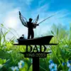 Black metal lawn memorial silhouette of an angelic fisherman in a boat with a fishing rod and the word DAD and dates 1946–2023 against a sunny grassy field with blue sky