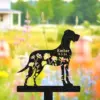 Black metal dog garden stake silhouette with floral cutouts and gold text reading "Ember 9.5.24" against a blurred colorful garden background