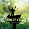 Black metal memorial silhouette of an angelic fisherman in a boat with 'DAD' and dates 1946–2023, shown among daisies and blurred green bokeh background