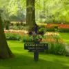253-1 Black memorial lawn cross with praying hands and rosary reading "In Loving Memory of Amaln Robinson 1962 - 2025" placed in a grassy garden with tulip beds