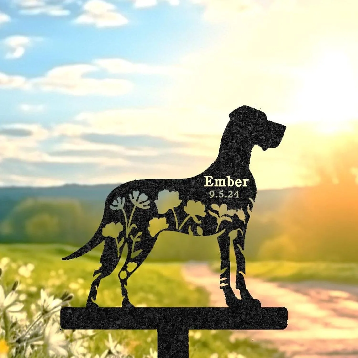 Black metal dog silhouette garden stake with floral cutouts and text "Ember 9.5.24" against a sunlit meadow background