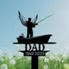 Silhouette memorial stake of a man with angel wings fishing from a boat above the word DAD and dates 1946-2023 against a blue sky and dandelion field