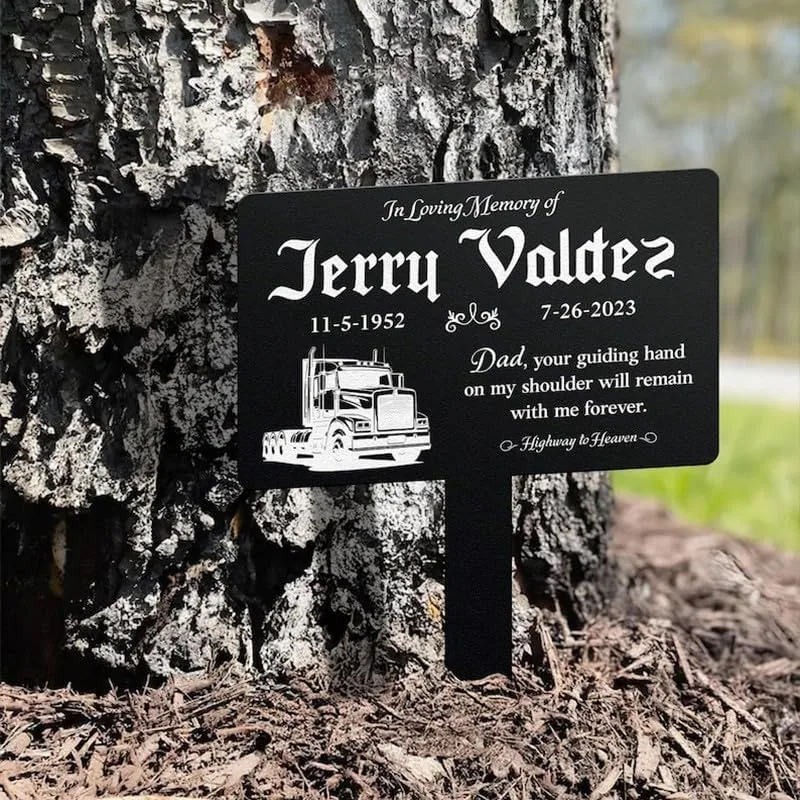 Black memorial stake beside a tree reading "In Loving Memory of Jerry Valdez 11-5-1952 7-26-2023" with a truck illustration and the inscription "Dad, your guiding hand on my shoulder will remain with me forever."