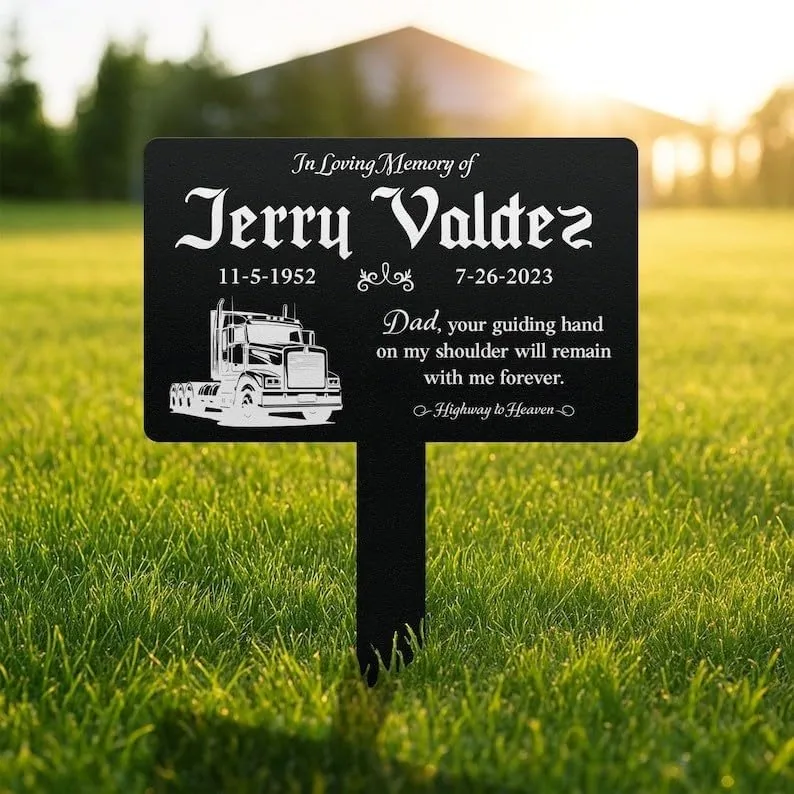 Black lawn memorial sign reading ‘In Loving Memory of Jerry Valdez 11-5-1952 7-26-2023’ with a truck illustration and the inscription ‘Dad, your guiding hand on my shoulder will remain with me forever,’ placed on grass outdoors