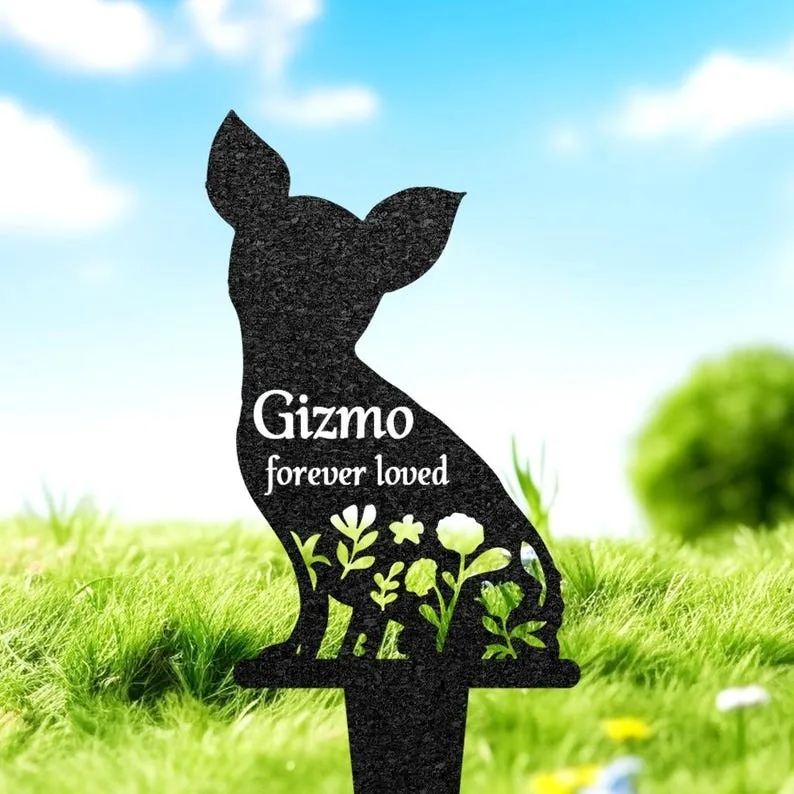 Black metal silhouette garden stake shaped like a small dog with cutout flowers and the text "Gizmo forever loved" displayed, placed in grassy lawn under a blue sky
