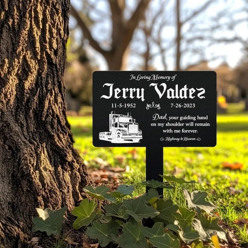 Black memorial lawn sign reading "In Loving Memory of Jerry Valdez 11-5-1952 - 7-26-2023" with a truck illustration and inscription "Dad, your guiding hand on my shoulder will remain with me forever," staked beside a tree in a sunlit yard